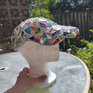 May include: A mosaic baseball cap with a white base and a visor adorned with colorful glass pieces. The cap is displayed on a white mannequin head, showcasing the intricate design and craftsmanship. The glass pieces are in various shapes and colors.