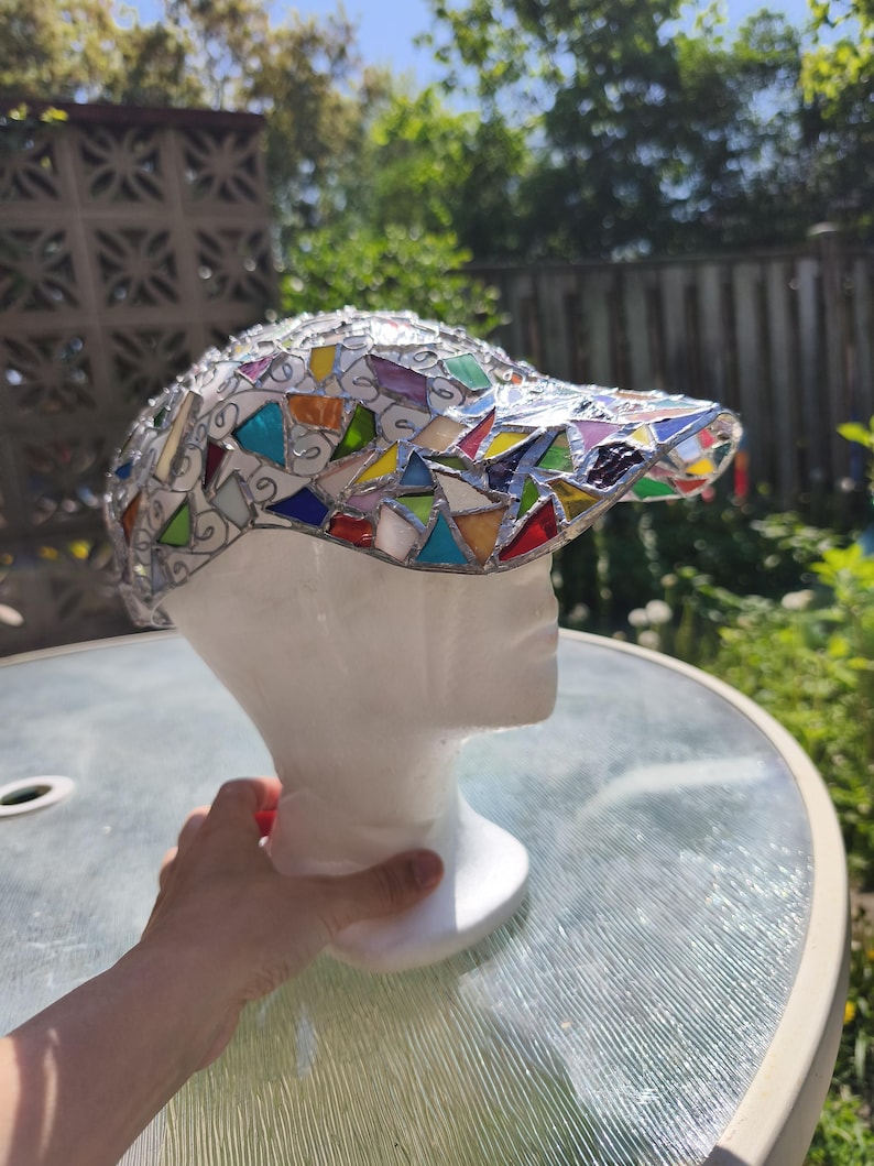 May include: A mosaic baseball cap with a white base and a visor adorned with colorful glass pieces. The cap is displayed on a white mannequin head, showcasing the intricate design and craftsmanship. The glass pieces are in various shapes and colors.