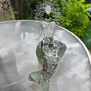 Puede incluir: Una escultura decorativa de sirena hecha de alambre plateado y piezas de vidrio marino esmerilado. La figura presenta una cabeza y un torso formados por alambre con detalles de vidrio blanco y una cola hecha de vidrio marino verde.