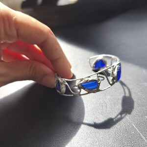 May include: A silver-toned cuff bracelet with blue glass inserts. The bracelet features an open design with decorative swirls and leaf-like shapes. The blue glass pieces are rectangular and oval, adding a pop of color to the jewelry.