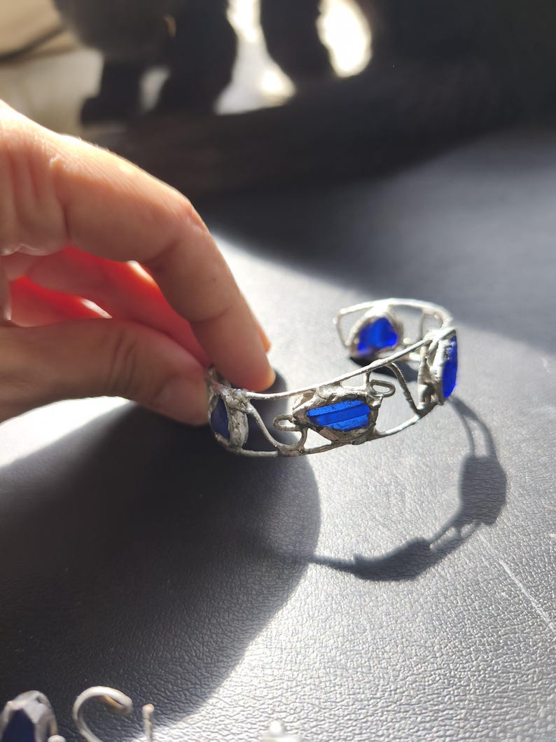 May include: A silver-toned cuff bracelet with blue glass inserts. The bracelet features an open design with decorative swirls and leaf-like shapes. The blue glass pieces are rectangular and oval, adding a pop of color to the jewelry.
