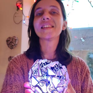May include: A woman holds a silver and white decorative sphere with a light inside. The sphere is made of many small pieces of glass and metal, and it is lit up with colorful lights. The woman is smiling and looking at the camera.