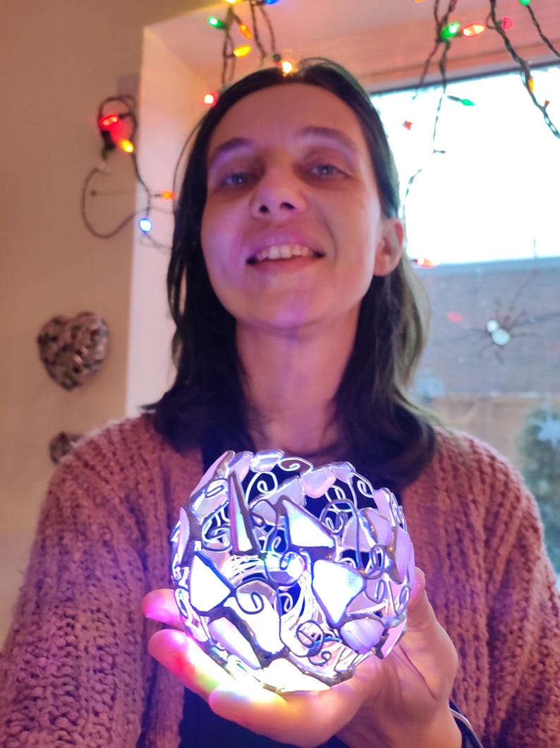 May include: A woman holds a silver and white decorative sphere with a light inside. The sphere is made of many small pieces of glass and metal, and it is lit up with colorful lights. The woman is smiling and looking at the camera.