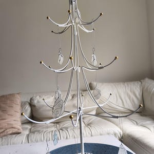 May include: A white metal Christmas tree-shaped ornament holder with gold accents and a gold star on top. The tree holds several clear, wire-wrapped ornaments. The tree is on a table with a blue placemat.