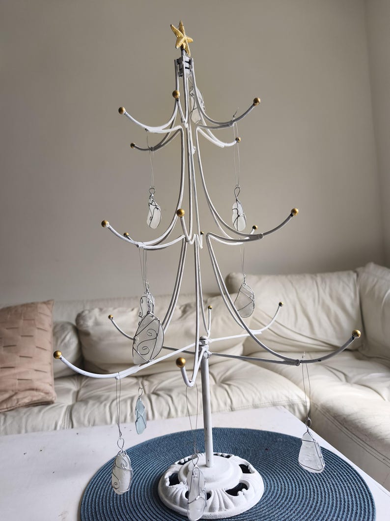 May include: A white metal Christmas tree-shaped ornament holder with gold accents and a gold star on top. The tree holds several clear, wire-wrapped ornaments. The tree is on a table with a blue placemat.