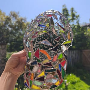 May include: A colorful stained glass vase with a mosaic design. The vase is composed of various geometric shapes in shades of red, yellow, green, blue, and orange, held together by silver-colored metal. The vase is held up against a sunny background.