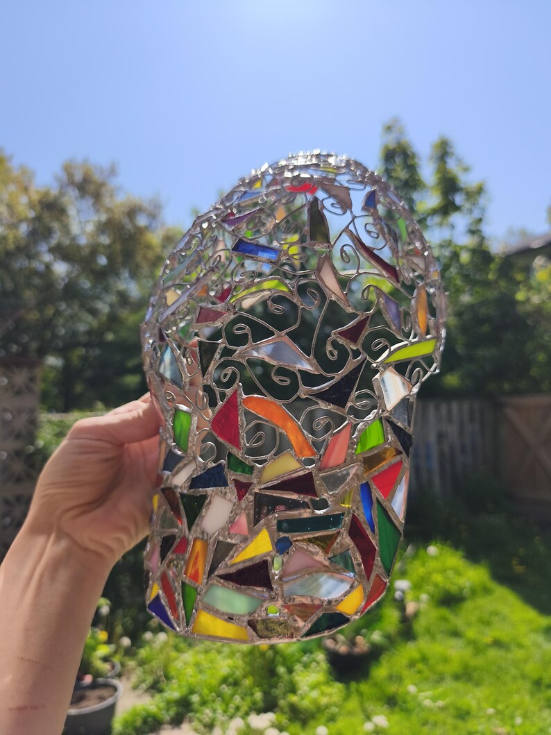 May include: A colorful stained glass vase with a mosaic design. The vase is composed of various geometric shapes in shades of red, yellow, green, blue, and orange, held together by silver-colored metal. The vase is held up against a sunny background.