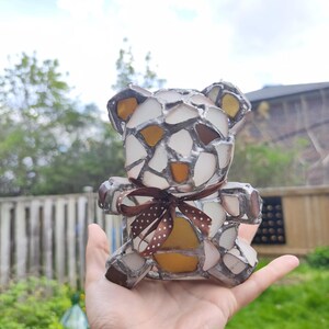 May include: A mosaic teddy bear figurine with a brown ribbon. The bear is made of white, brown, and yellow glass pieces. The bear is held in a hand against a background of green foliage and a cloudy sky.