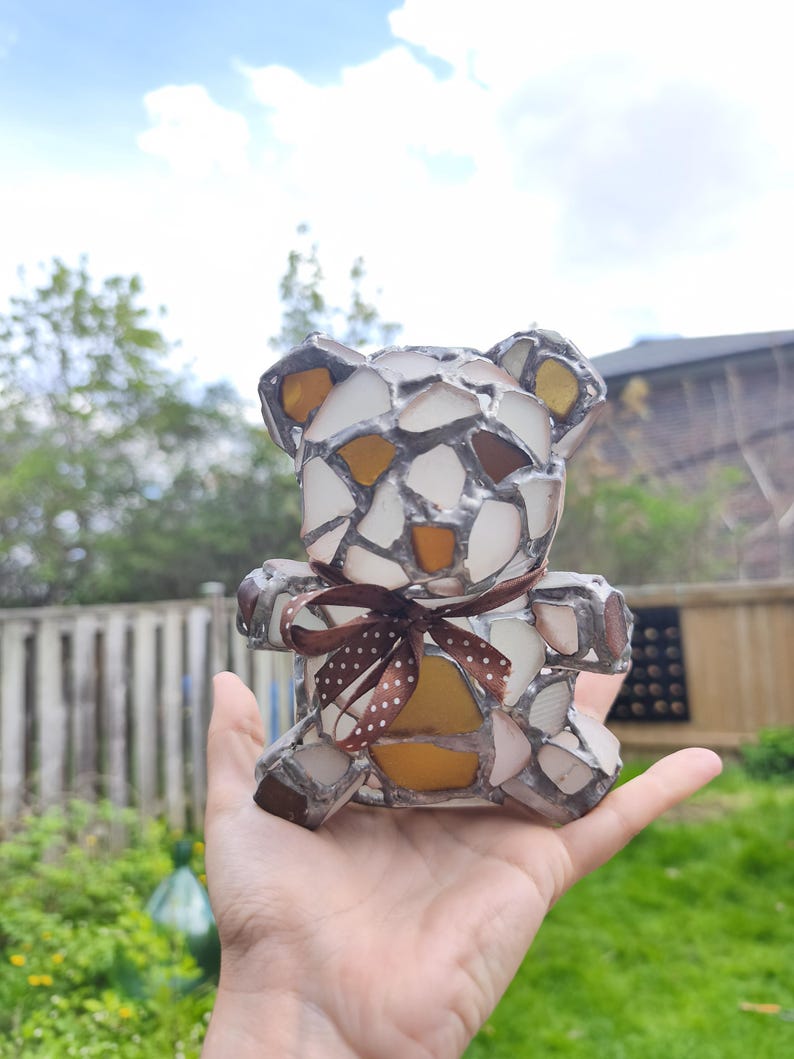 May include: A mosaic teddy bear figurine with a brown ribbon. The bear is made of white, brown, and yellow glass pieces. The bear is held in a hand against a background of green foliage and a cloudy sky.