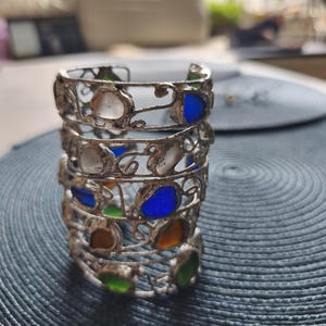 May include: A stack of silver-toned metal bracelets with colorful glass inserts. The bracelets feature a filigree design and are adorned with blue, green, orange, and white glass pieces. The bracelets are stacked on a textured blue surface.