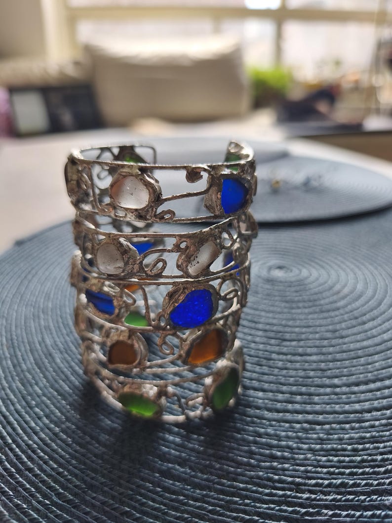 May include: A stack of silver-toned metal bracelets with colorful glass inserts. The bracelets feature a filigree design and are adorned with blue, green, orange, and white glass pieces. The bracelets are stacked on a textured blue surface.