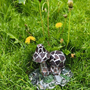 May include: A handcrafted glass mushroom sculpture with two mushroom caps. The caps are brown and silver, and the stems are clear. The sculpture sits on a base of green and gray glass pieces, surrounded by green grass and dandelions.