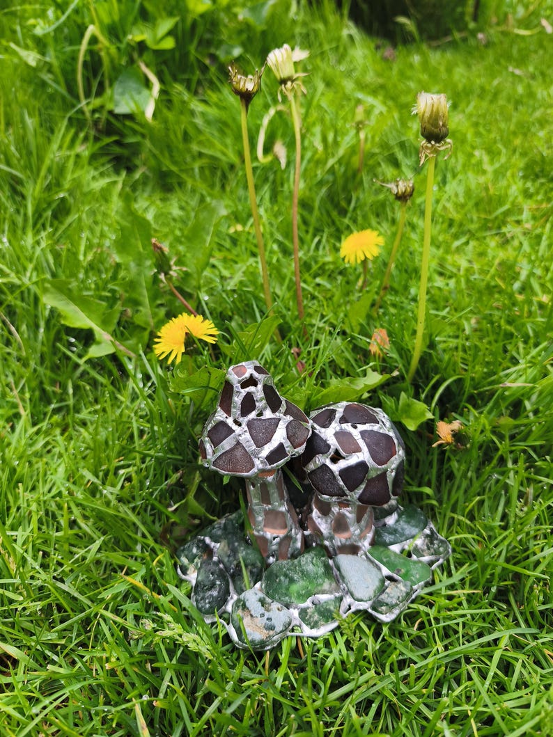 May include: A handcrafted glass mushroom sculpture with two mushroom caps. The caps are brown and silver, and the stems are clear. The sculpture sits on a base of green and gray glass pieces, surrounded by green grass and dandelions.