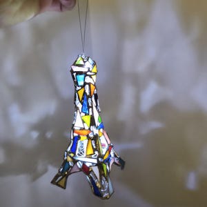 May include: A stained glass Eiffel Tower ornament in a mosaic of colours. The ornament is made of various coloured glass pieces, including blue, yellow, orange, and white. It is suspended by a thin wire, and the light shines through the glass.