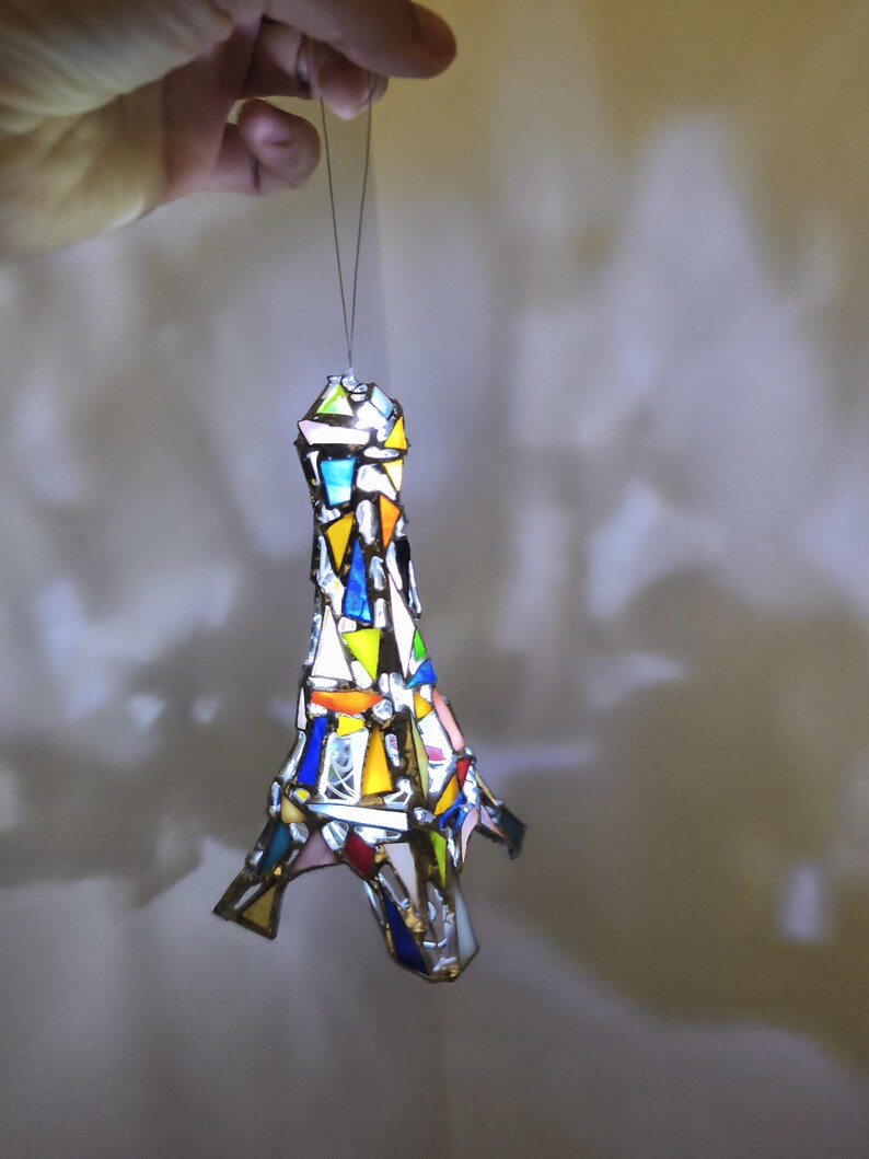 May include: A stained glass Eiffel Tower ornament in a mosaic of colours. The ornament is made of various coloured glass pieces, including blue, yellow, orange, and white. It is suspended by a thin wire, and the light shines through the glass.
