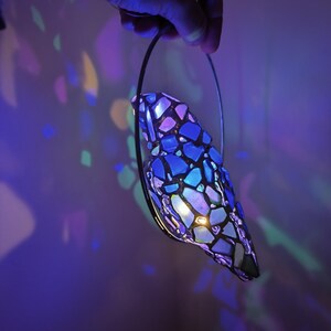 May include: A stained glass lamp with a mosaic design in shades of blue, purple, and white is held by a hand. The lamp is illuminated, casting colorful light patterns on the wall. The lamp has a black metal frame.