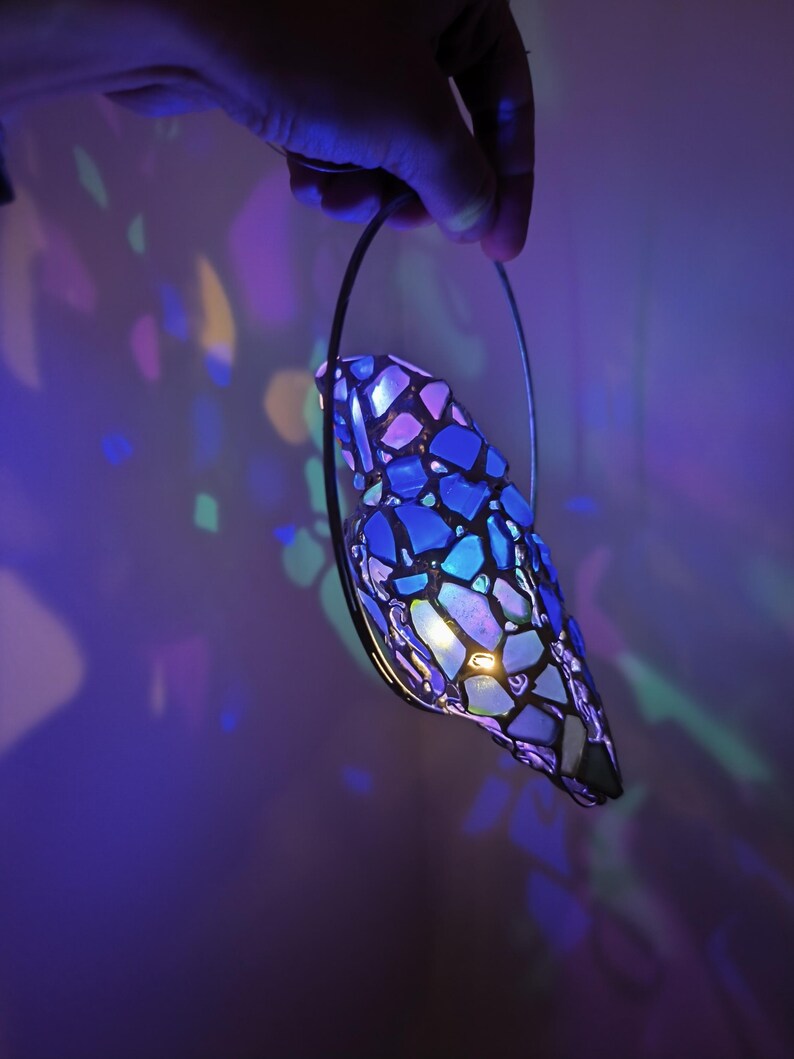 May include: A stained glass lamp with a mosaic design in shades of blue, purple, and white is held by a hand. The lamp is illuminated, casting colorful light patterns on the wall. The lamp has a black metal frame.