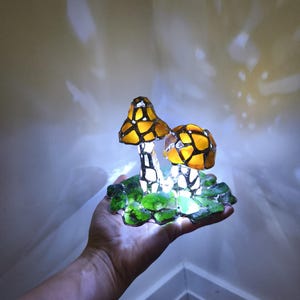 May include: A decorative mushroom lamp with two illuminated, stained-glass style mushroom caps in yellow and orange. The mushrooms sit on a base of green glass pieces. The lamp is held in a hand.