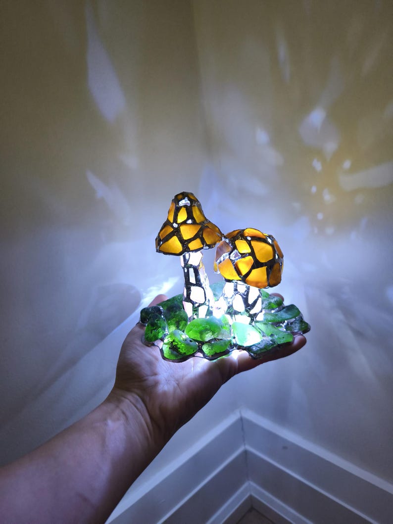 May include: A decorative mushroom lamp with two illuminated, stained-glass style mushroom caps in yellow and orange. The mushrooms sit on a base of green glass pieces. The lamp is held in a hand.