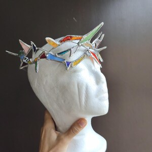May include: A crown constructed from multiple pieces of multicolored stained glass held together by silver solder. The crown features sharp, pointed segments in shades of red, blue, yellow, green, and white, arranged in a circular, jagged formation.