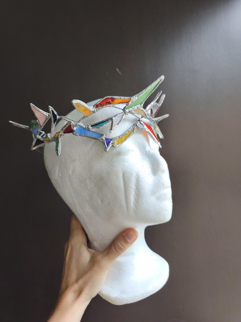 May include: A crown constructed from multiple pieces of multicolored stained glass held together by silver solder. The crown features sharp, pointed segments in shades of red, blue, yellow, green, and white, arranged in a circular, jagged formation.