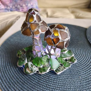 May include: A decorative glass mushroom sculpture with brown and amber caps, set on a base of green glass leaves. The mushrooms are made of various glass pieces, creating a mosaic effect. The sculpture sits on a blue placemat.