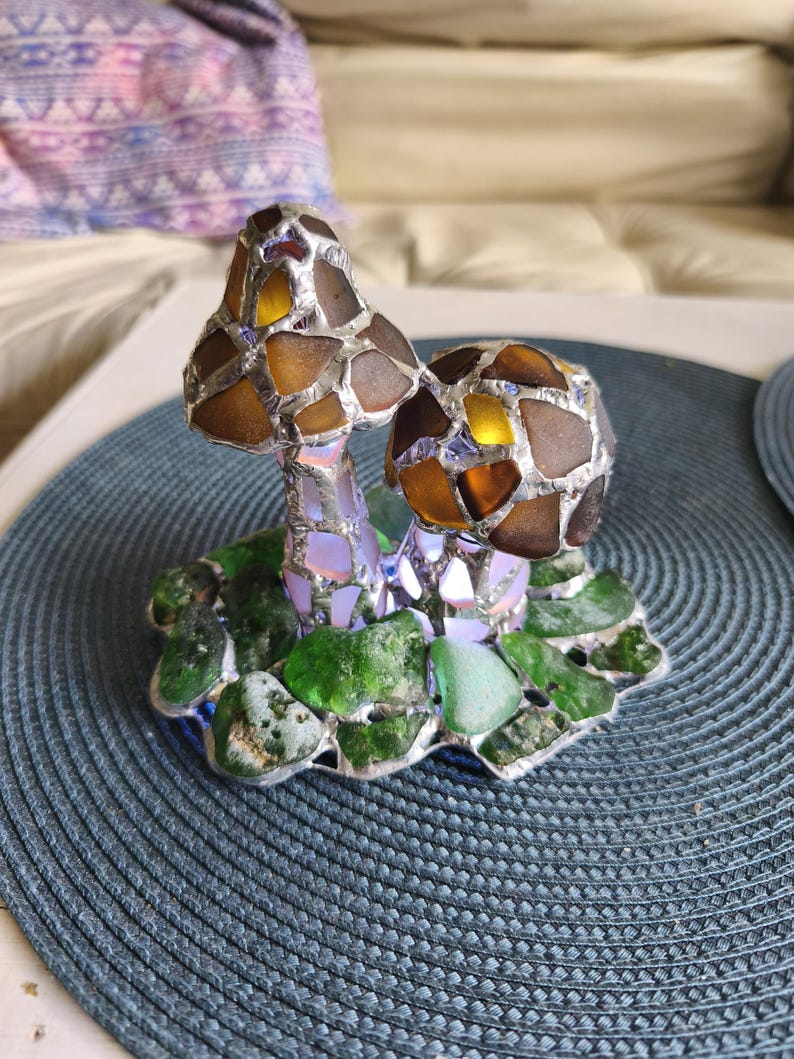 May include: A decorative glass mushroom sculpture with brown and amber caps, set on a base of green glass leaves. The mushrooms are made of various glass pieces, creating a mosaic effect. The sculpture sits on a blue placemat.
