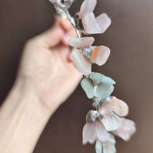 May include: A handcrafted floral hair accessory featuring a silver-toned wire base adorned with sea glass in shades of pink, peach, and pale green. The sea glass pieces are shaped like petals, creating a delicate and natural look. This unique piece is perfect for weddings or special occasions.