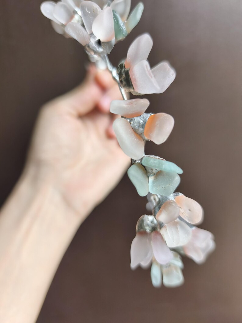 May include: A handcrafted floral hair accessory featuring a silver-toned wire base adorned with sea glass in shades of pink, peach, and pale green. The sea glass pieces are shaped like petals, creating a delicate and natural look. This unique piece is perfect for weddings or special occasions.