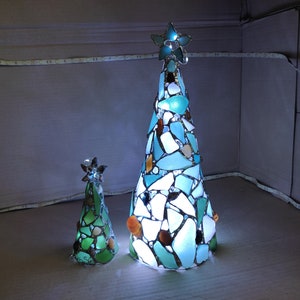 Pode incluir: Duas decora&ccedil;&otilde;es de &aacute;rvore de Natal iluminadas. A &aacute;rvore maior tem formato c&ocirc;nico, adornada com pe&ccedil;as de vidro azuis, brancas e marrons, e encimada por uma estrela. A &aacute;rvore menor &eacute; verde e tamb&eacute;m tem uma estrela.