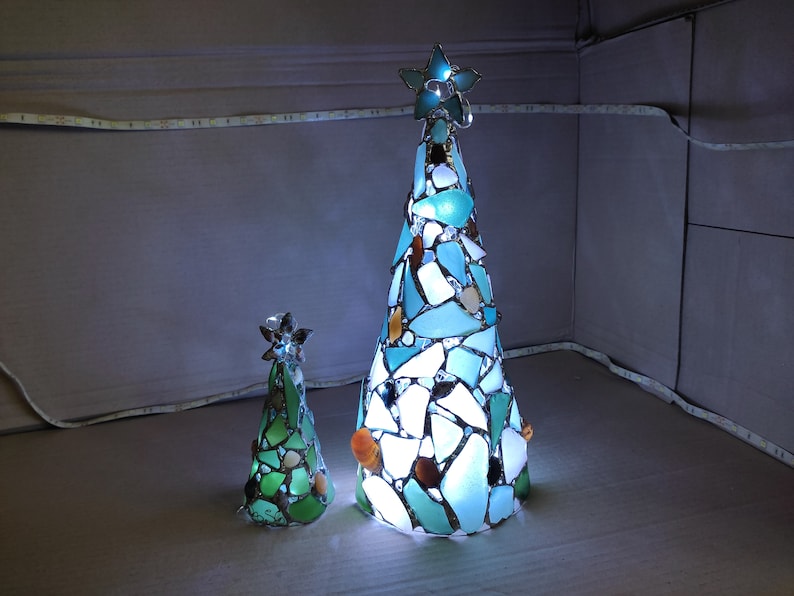 Pode incluir: Duas decora&ccedil;&otilde;es de &aacute;rvore de Natal iluminadas. A &aacute;rvore maior tem formato c&ocirc;nico, adornada com pe&ccedil;as de vidro azuis, brancas e marrons, e encimada por uma estrela. A &aacute;rvore menor &eacute; verde e tamb&eacute;m tem uma estrela.
