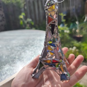 May include: A stained glass Eiffel Tower ornament, featuring a mosaic of coloured glass pieces. The glass is in shades of red, yellow, blue, and green, held together with silver solder. A white string is attached for hanging. A decorative item.