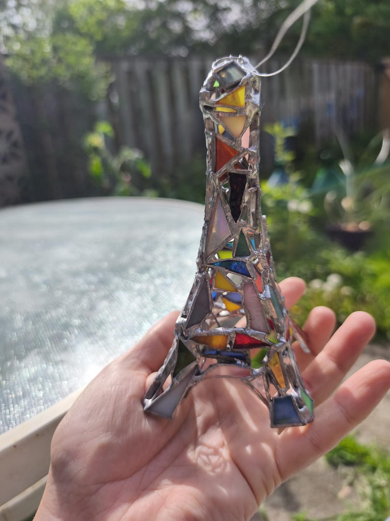 May include: A stained glass Eiffel Tower ornament, featuring a mosaic of coloured glass pieces. The glass is in shades of red, yellow, blue, and green, held together with silver solder. A white string is attached for hanging. A decorative item.