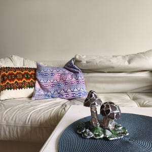 May include: A decorative glass mushroom sculpture with brown and silver accents sits on a blue placemat. The sculpture has two mushrooms with textured caps and sits on a green base. Pillows with colorful patterns are on a white couch.