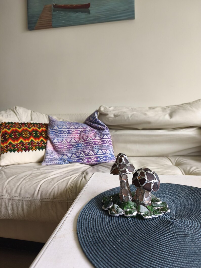 May include: A decorative glass mushroom sculpture with brown and silver accents sits on a blue placemat. The sculpture has two mushrooms with textured caps and sits on a green base. Pillows with colorful patterns are on a white couch.