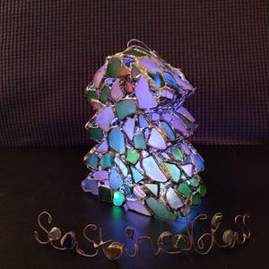 K&ouml;nnte beinhalten: Eine Glaslampe aus Meerglas in Gr&uuml;n-, Blau- und Wei&szlig;t&ouml;nen. Die Lampe ist beleuchtet und strahlt ein buntes Licht ab. Die Worte "Sea Stones" sind in Draht unter der Lampe geschrieben.