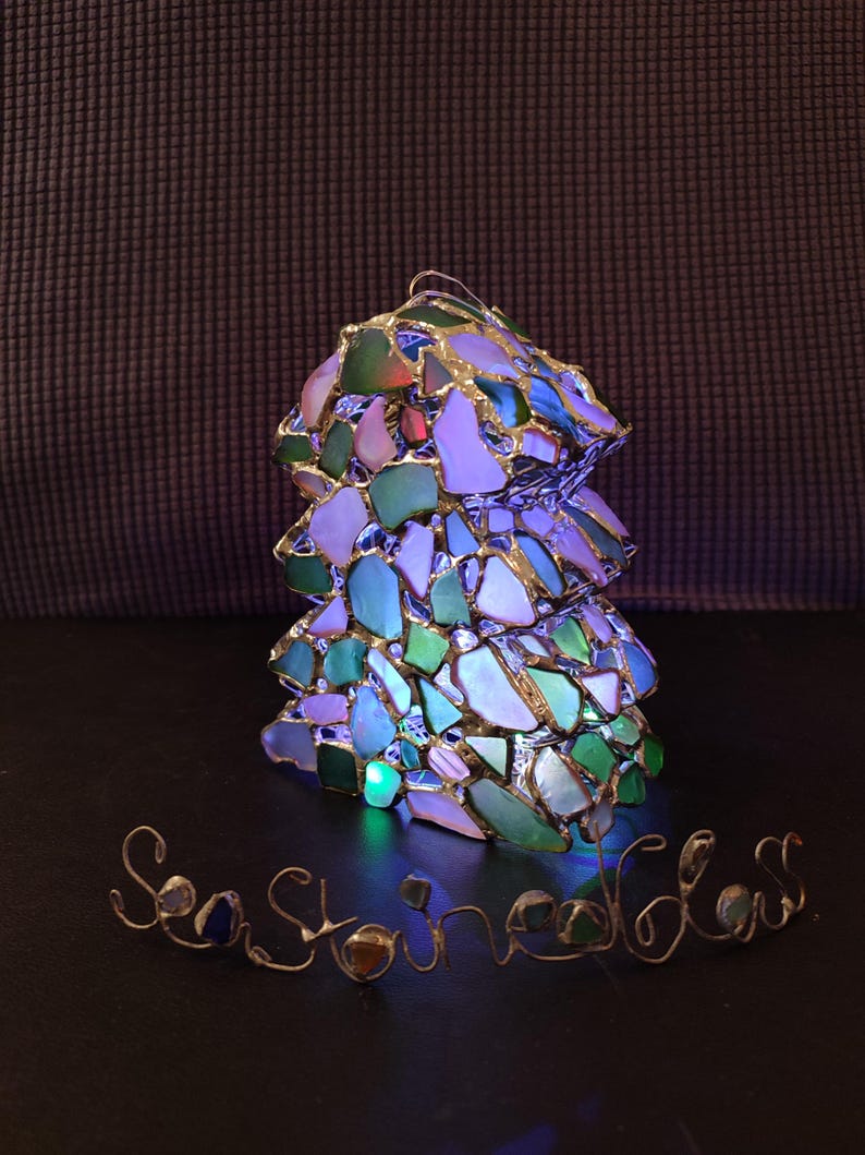 K&ouml;nnte beinhalten: Eine Glaslampe aus Meerglas in Gr&uuml;n-, Blau- und Wei&szlig;t&ouml;nen. Die Lampe ist beleuchtet und strahlt ein buntes Licht ab. Die Worte "Sea Stones" sind in Draht unter der Lampe geschrieben.