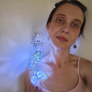 Puede incluir: Una figura decorativa de sirena hecha de alambre y piezas de vidrio azul transl&uacute;cido. La sirena presenta una cabeza y un torso formados por alambre con una cola construida con fragmentos de vidrio estilo mosaico que emiten una luz azul brillante.