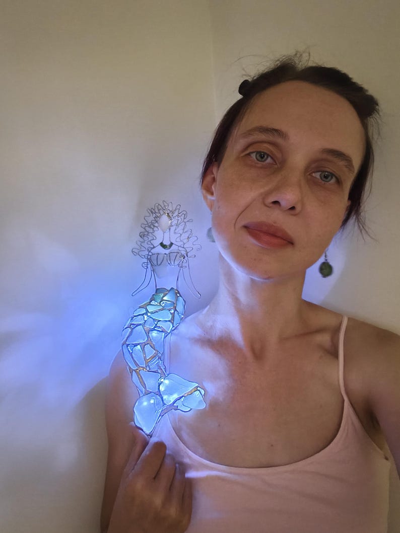 Puede incluir: Una figura decorativa de sirena hecha de alambre y piezas de vidrio azul transl&uacute;cido. La sirena presenta una cabeza y un torso formados por alambre con una cola construida con fragmentos de vidrio estilo mosaico que emiten una luz azul brillante.