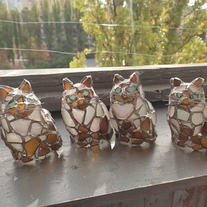 May include: Four stained glass cat figurines, each with a different pose and color variation. The cats are made of small pieces of glass in shades of white, brown, and green.