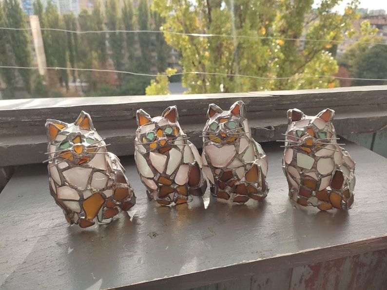May include: Four stained glass cat figurines, each with a different pose and color variation. The cats are made of small pieces of glass in shades of white, brown, and green.