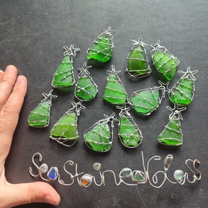 May include: A collection of 13 green glass Christmas tree ornaments wrapped in silver wire. The ornaments are arranged in a circle on a black surface. The words "Sea Glass" are written in silver wire below the ornaments.