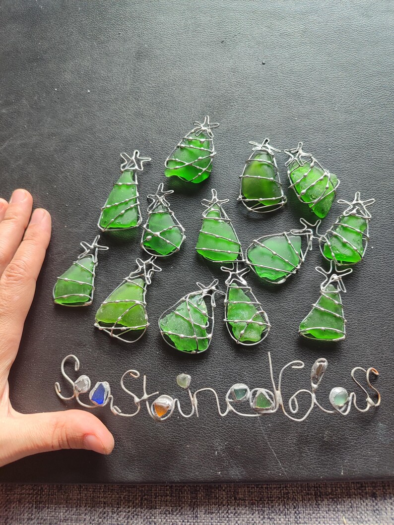 May include: A collection of 13 green glass Christmas tree ornaments wrapped in silver wire. The ornaments are arranged in a circle on a black surface. The words "Sea Glass" are written in silver wire below the ornaments.