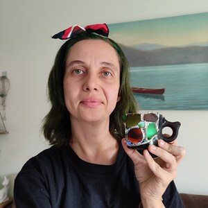 May include: A woman with green hair is holding a small, colorful mosaic mug with a black handle. The mug is made of small pieces of glass and has a variety of colors, including green, blue, brown, and red.