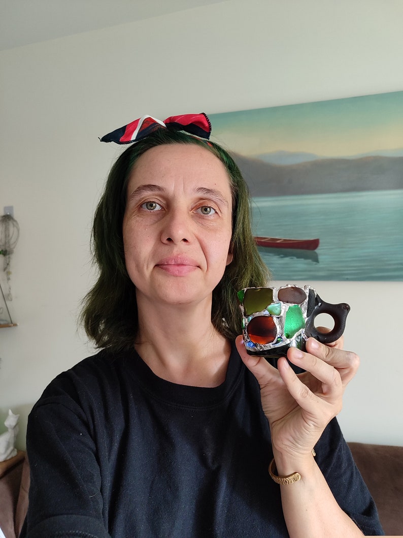 May include: A woman with green hair is holding a small, colorful mosaic mug with a black handle. The mug is made of small pieces of glass and has a variety of colors, including green, blue, brown, and red.