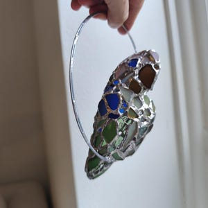 May include: A stained glass heart-shaped ornament with a silver metal ring for hanging. The heart is composed of various colors of glass, including blue, green, brown, and clear. The ornament is held up by a hand.