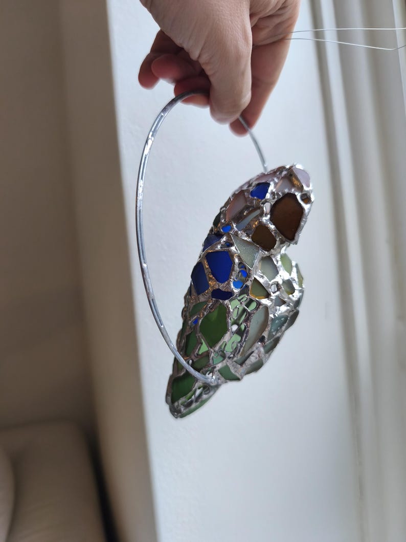 May include: A stained glass heart-shaped ornament with a silver metal ring for hanging. The heart is composed of various colors of glass, including blue, green, brown, and clear. The ornament is held up by a hand.