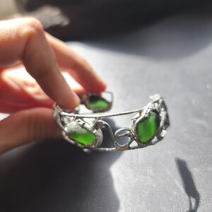 May include: A silver-toned bracelet featuring three vibrant green sea glass stones. The bracelet has an open design with decorative filigree details. The sea glass is a bright, translucent green color.