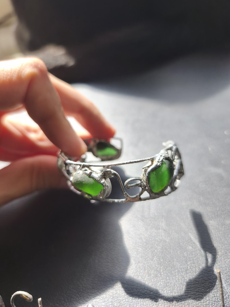 May include: A silver-toned bracelet featuring three vibrant green sea glass stones. The bracelet has an open design with decorative filigree details. The sea glass is a bright, translucent green color.