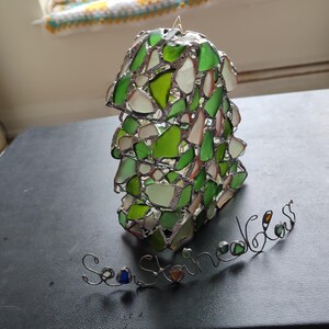 K&ouml;nnte beinhalten: Ein gr&uuml;ner und wei&szlig;er Meerglassonnenf&auml;nger mit den Worten "Sea Shine Glass" in Draht.